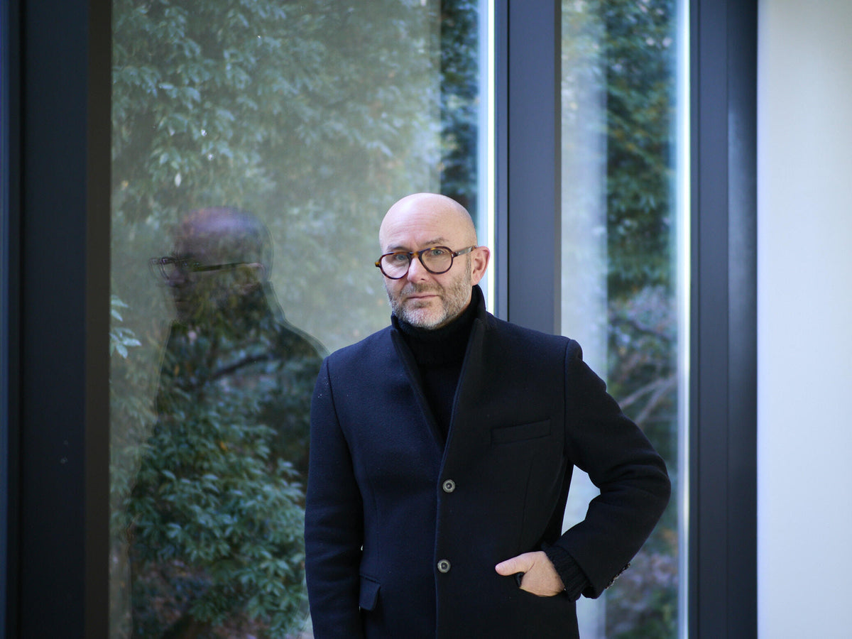 Portrait of Solve Sundsbo wearing round glasses and a dark coat, standing beside large windows with greenery visible outside.
