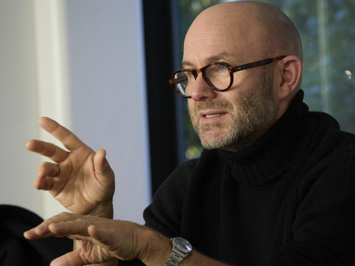 Solve Sundsbo wearing round glasses, a black turtleneck, and a dark coat, standing beside large windows with his reflection visible in the glass.