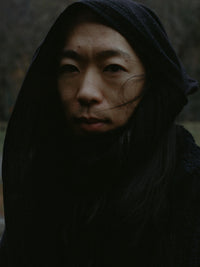 Close-up portrait of a woman with long dark hair wrapped in a black hood, looking toward the camera.