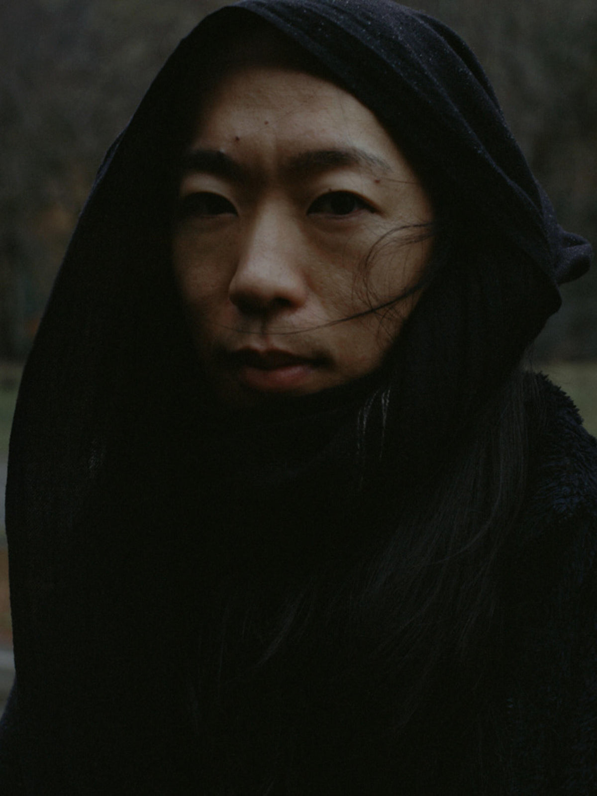 Close-up portrait of a woman with long dark hair wrapped in a black hood, looking toward the camera.