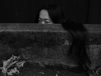 Black-and-white portrait of a woman peering over a concrete wall, with her long hair cascading down its side.