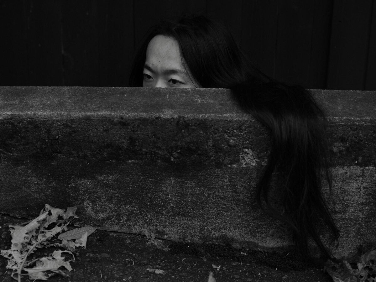 Black-and-white portrait of a woman peering over a concrete wall, with her long hair cascading down its side.