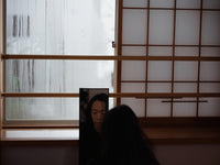 Woman with long dark hair facing a small mirror inside a traditional Japanese-style room with shoji screens.