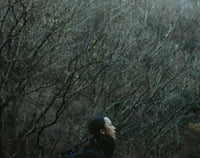 Woman in profile standing among dense, leafless trees in a dark woodland.
