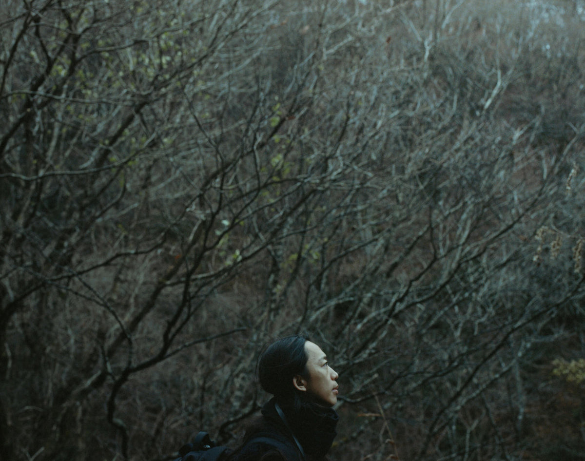 Woman in profile standing among dense, leafless trees in a dark woodland.