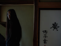 Woman with very long dark hair standing in a dim traditional interior beside a panel of Japanese calligraphy.