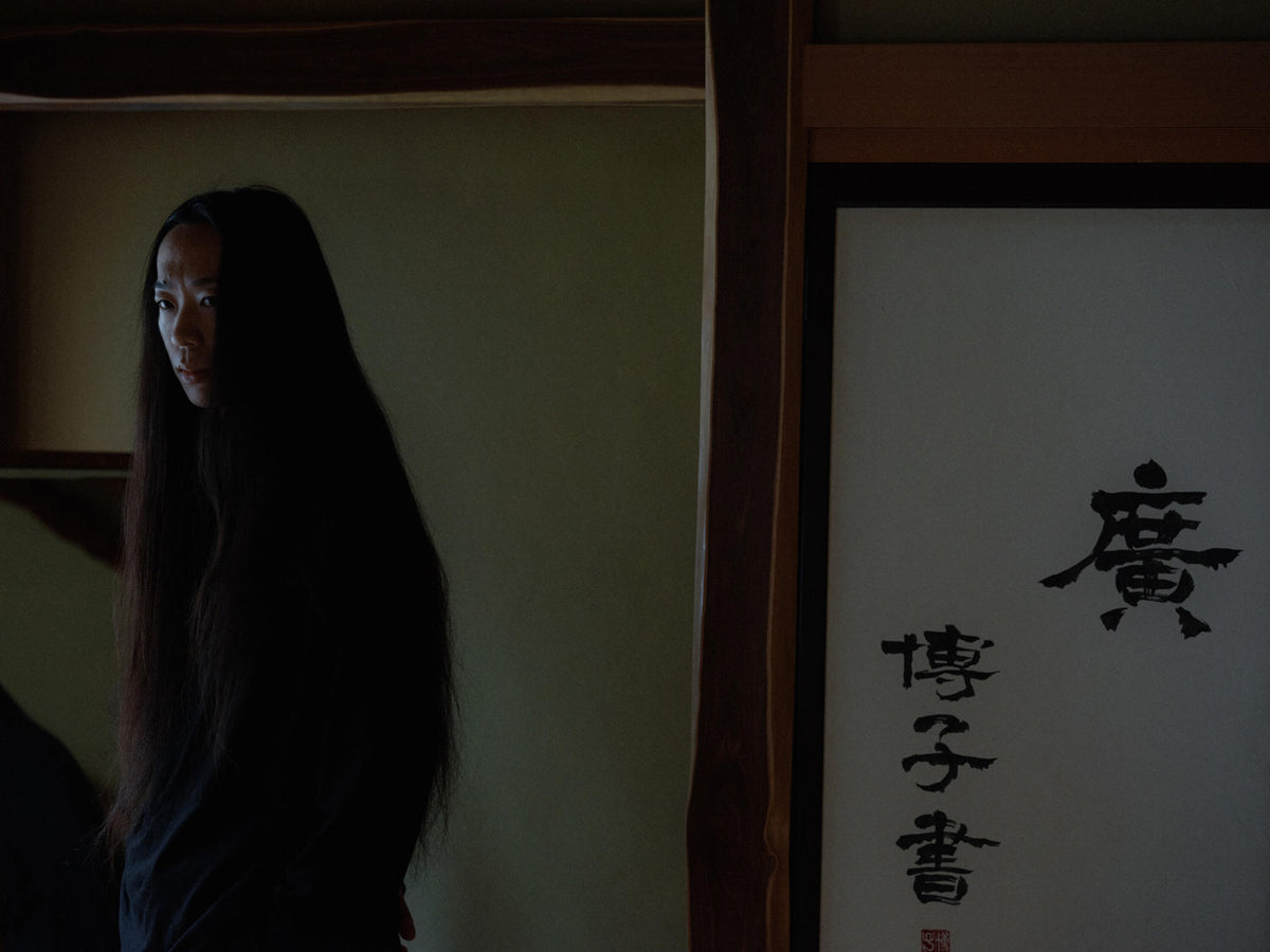 Woman with very long dark hair standing in a dim traditional interior beside a panel of Japanese calligraphy.