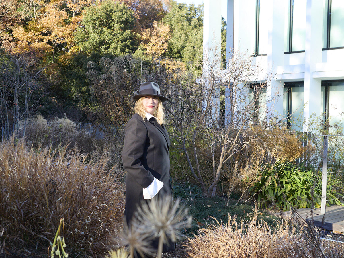 Portrait of Julia Hetta wearing a dark hat and long black coat, standing in a sunlit garden beside a modern building.