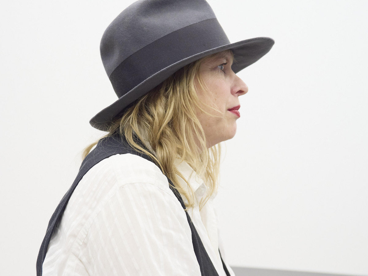 Side-profile portrait of a Julia Hetta wearing a wide-brimmed dark hat, white shirt, and dark vest against a white background.