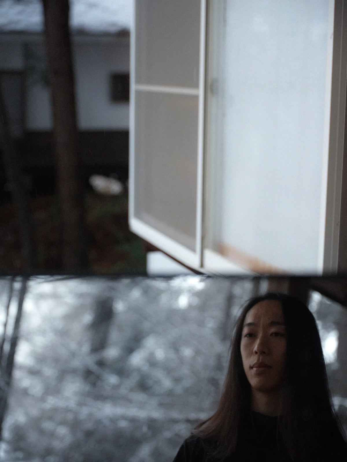 Portrait of a woman with long dark hair standing beneath a suspended architectural structure, with a blurred wooded landscape in the background.