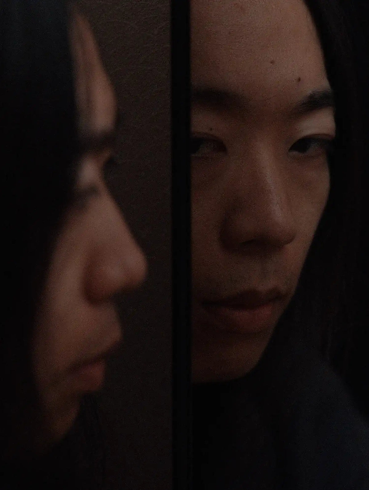 Close up portrait of a woman seen through a narrow mirror, with her face and reflection emerging from the darkness.