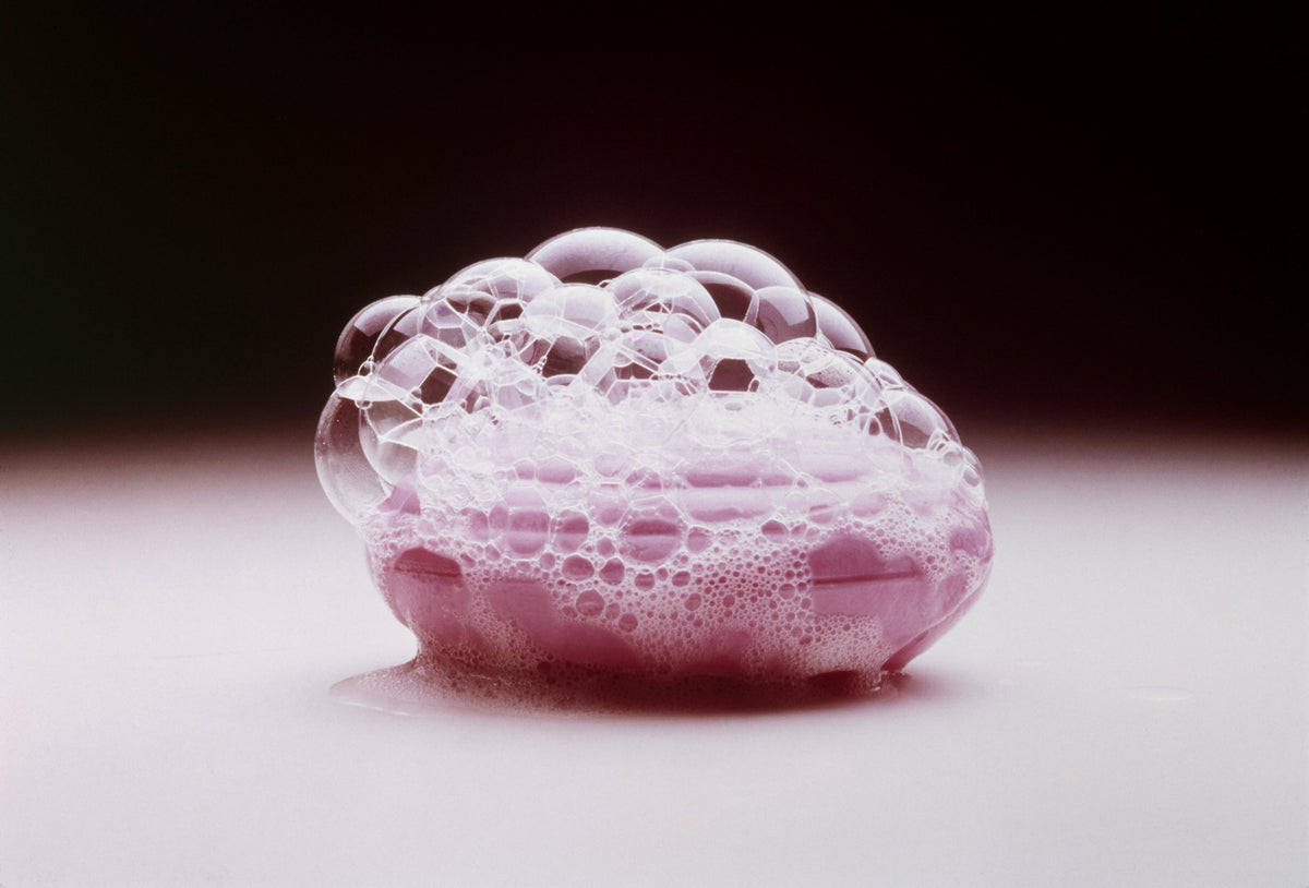 Composizione di bolle di sapone trasparenti e schiuma rosa, fotografata su una superficie chiara con sfondo scuro.